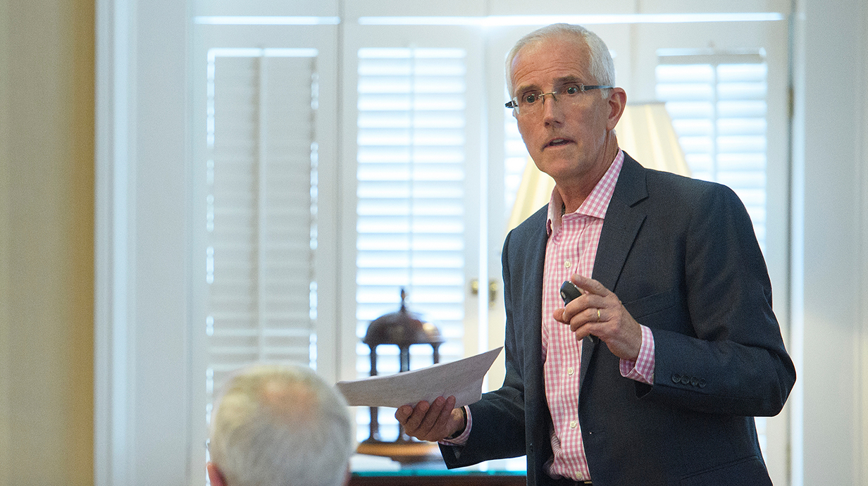 A man wearing glasses holds a remote control during a presentation in a meeting room.