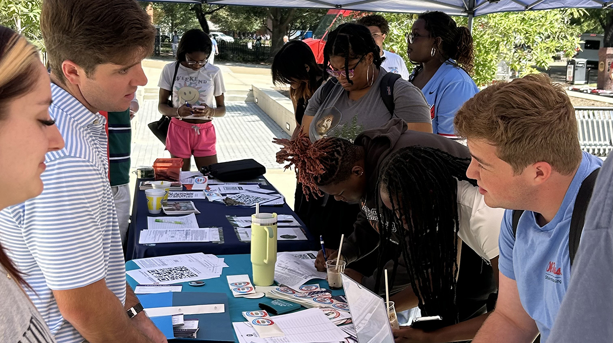 A group of young men and women fill out paperwork underneath a tent at an outdoor registration fair.