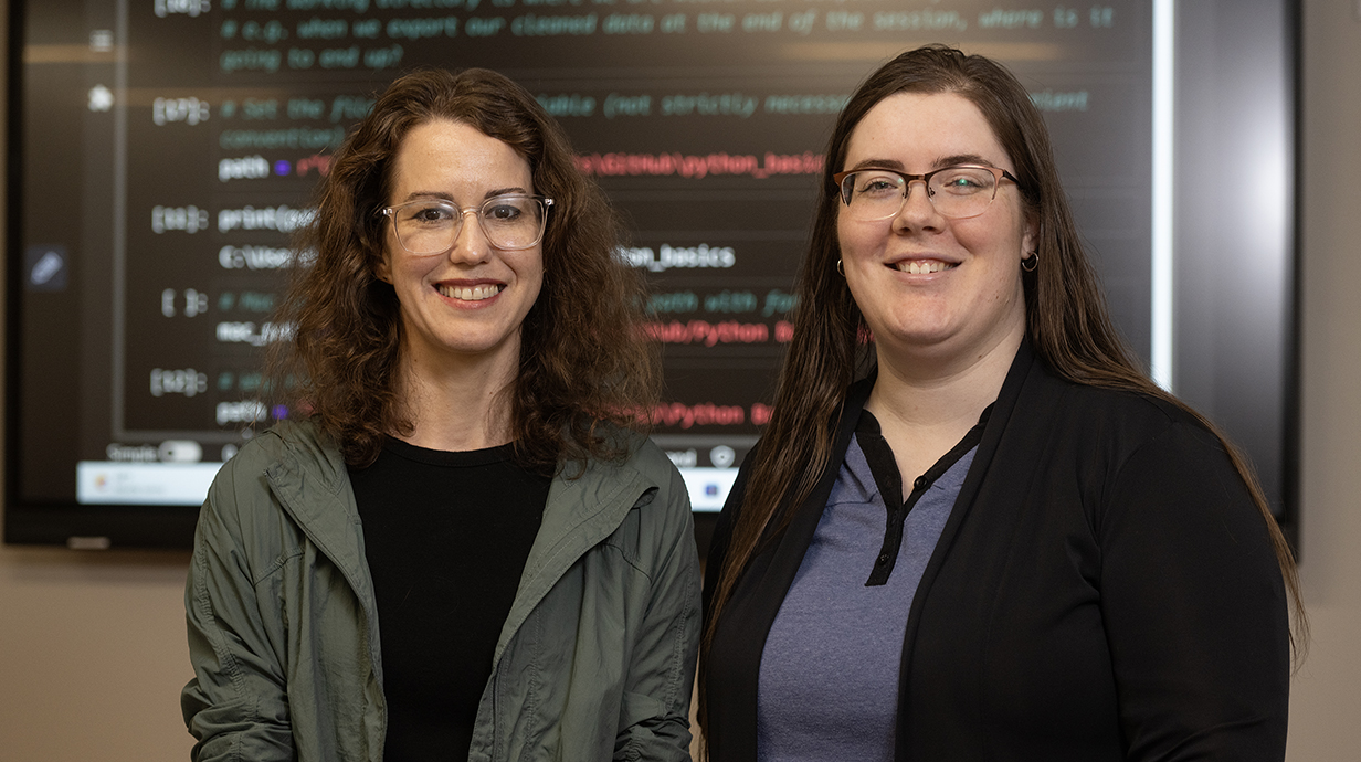 Two women stand in front of a video screen displaying computer code.