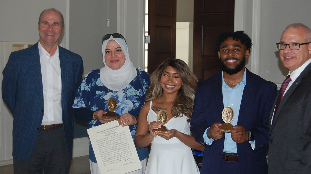 Three men and two women stand in a banquet hall; the women and one of the men hold gold awards.