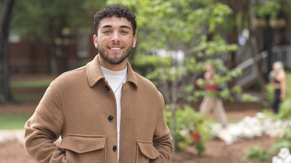 A young man wearing a suede coat stands in a park.