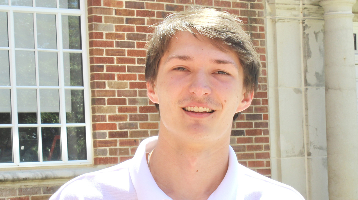 A young man wearing a white polo shirt poses in front of a brick building.