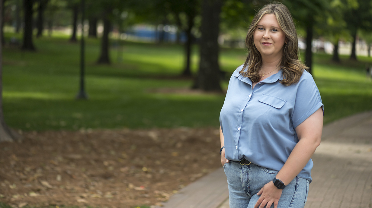 A woman wearing a light blue shirt and jeans stands smiling in a park.