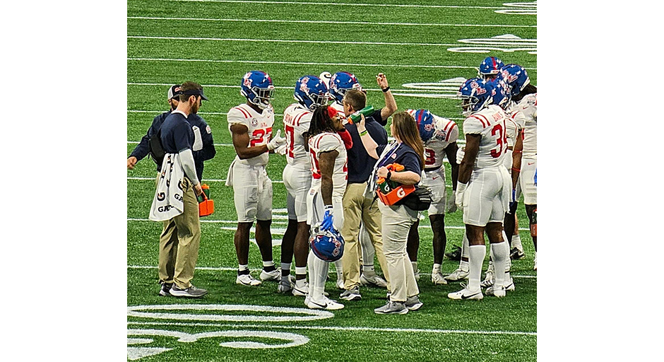 A young woman offers a water bottle to football players standing in a huddle.