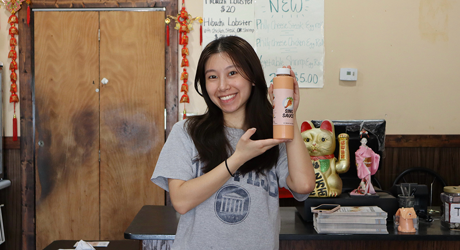 A young lady stands in a restaurant kitchen holding a bottle labeled Sing Sauce.