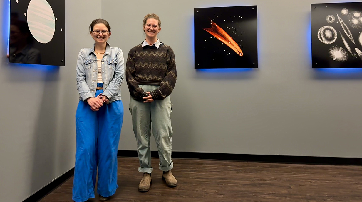 Two women stand in front of colorful museum displays of planets, comets and stars.
