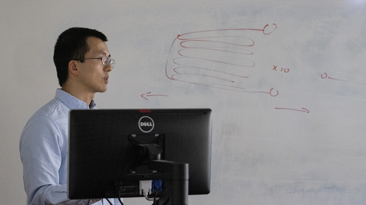 A amn wearing glasses stands in front of a whiteboard with an engineering diagram