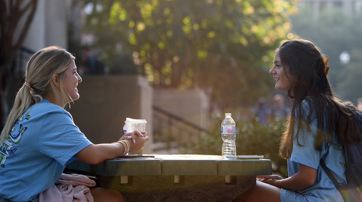 Two young women chat at a picnic table in a park.