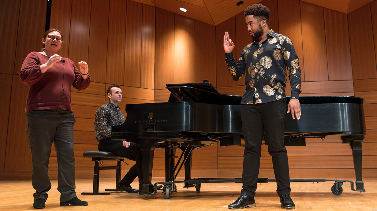 A woman gives direction to a man onstage while a pianist waits behind them at the keys.