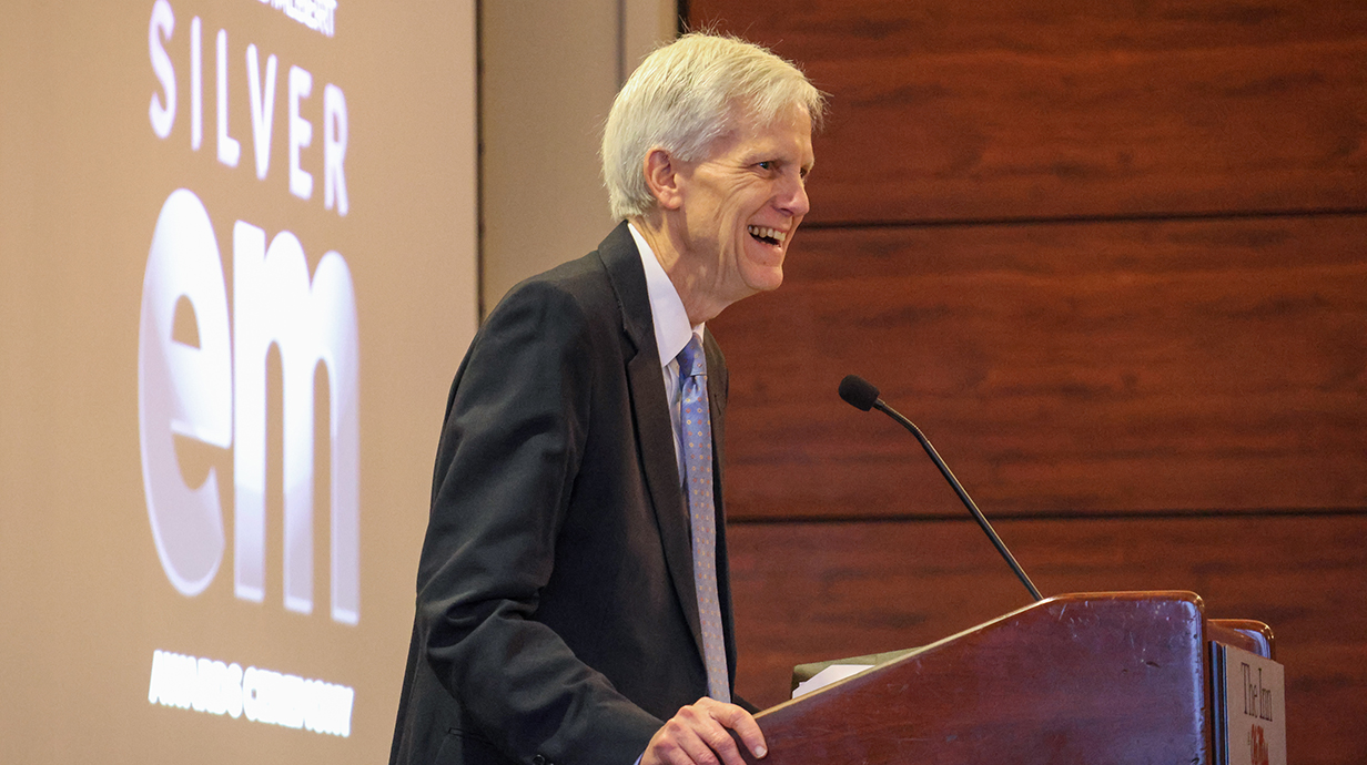 A man smiles while speaking at a podium in front of a projection screen.
