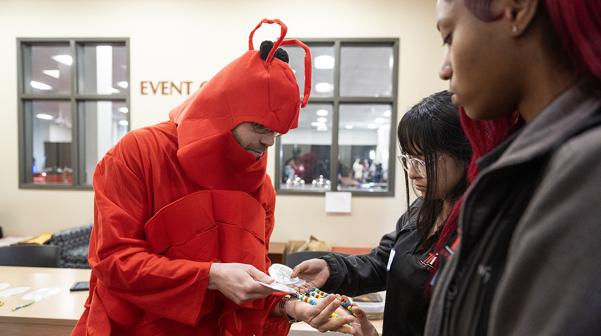 A man wearing a red crawfish costume shows a handful of beads to two students.