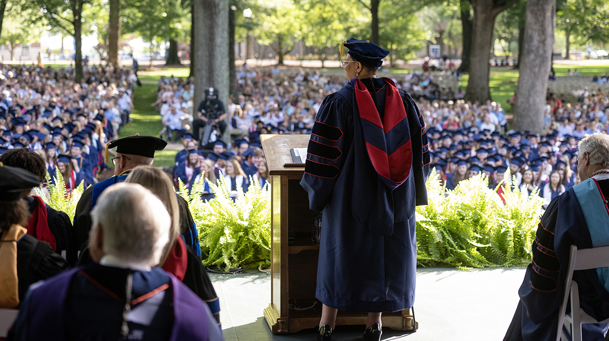A woman in graduation robes speaks at an outdoor graduation ceremony before a crowd of thousands of people.