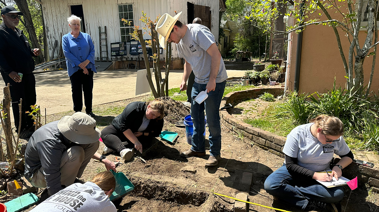 A young man wearing a cowboy hat stands at a dig site while other young people work close to the ground.