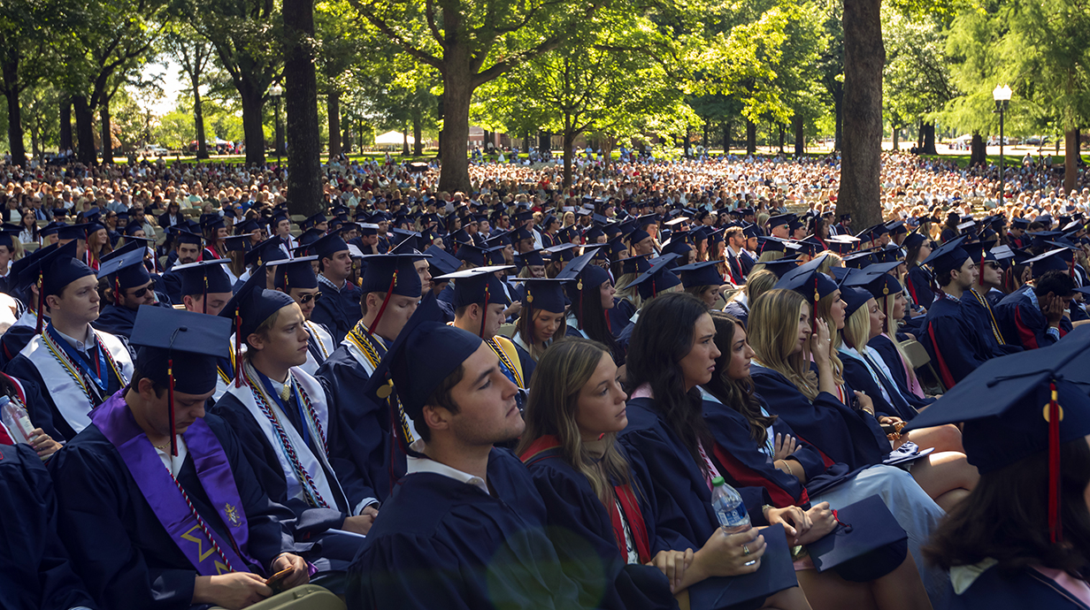 Thousands of people wearing graduation robes sit in a park on a sunny morning.