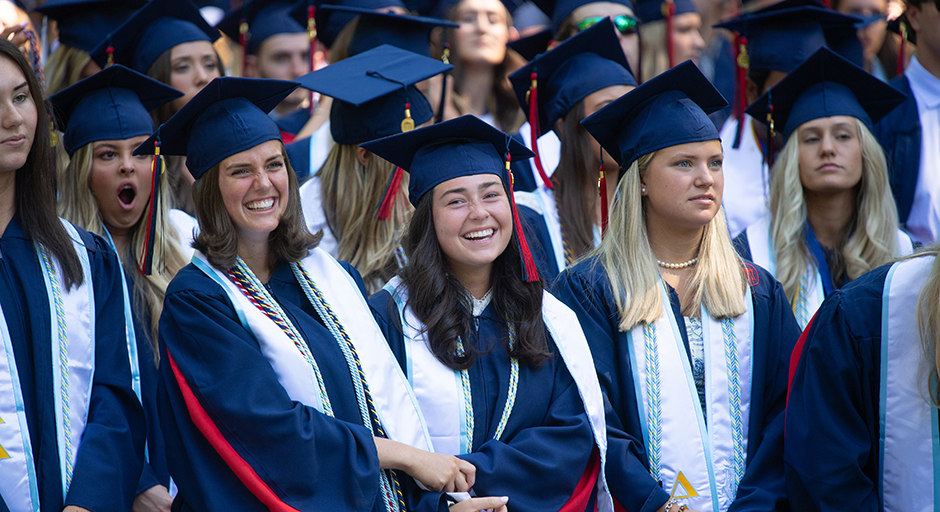 Young women wearing graduation regalia hold hands in a park.