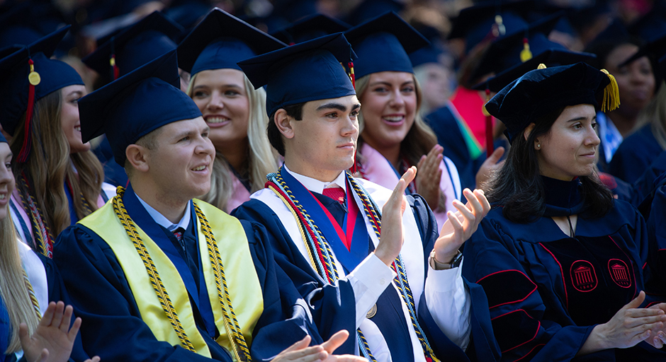 Two young men wearing robes applaud at a graduation ceremony.