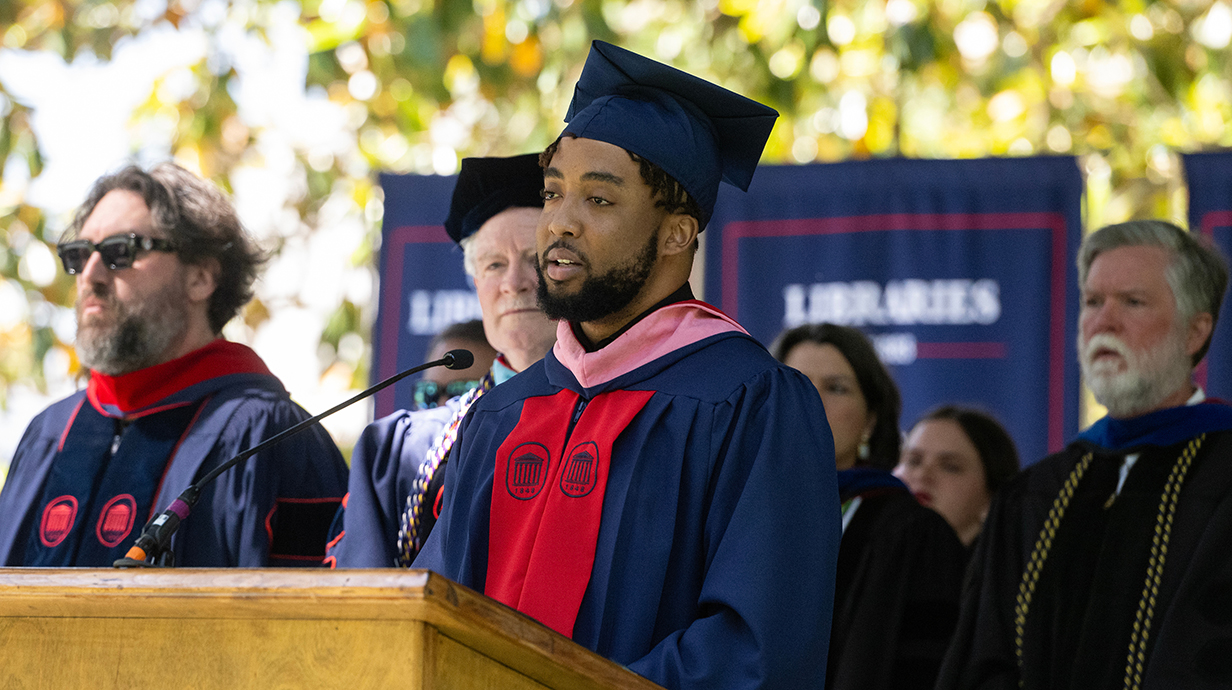A man wearing graduation regalia stands at a podium on an outdoor stage.