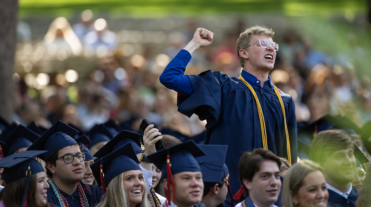 A young man stands and fist-pumps in celebration at a graduation ceremony.