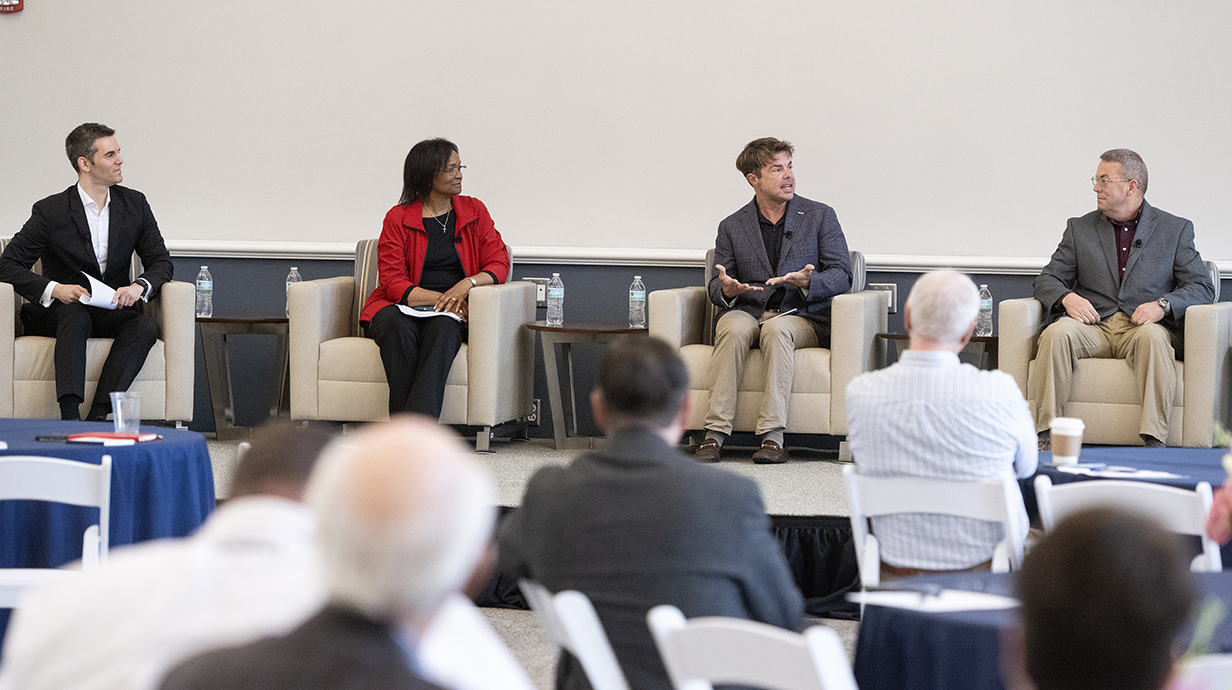 Three men and a woman talk on a stage at the front of a conference hall.