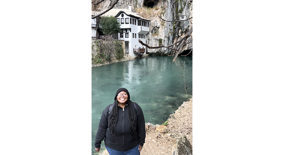 A young woman stands in front of a pool at the base of a cliff, across from a lakeside building.