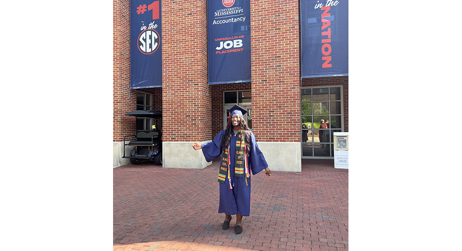 A young woman in graduation robes stands in front of a brick building.