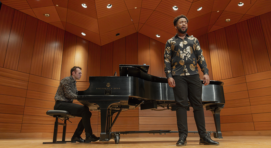 A man sings on a stage, accompanied by a pianist just behind him.