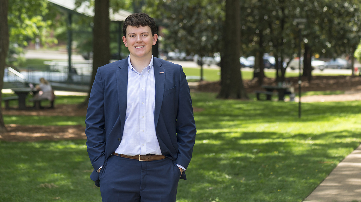 A young man in a blue sportcoat stands in a park.