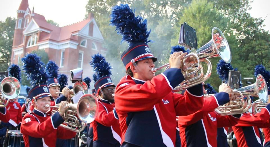 A marching band marches past a red building on a tree-lined street.