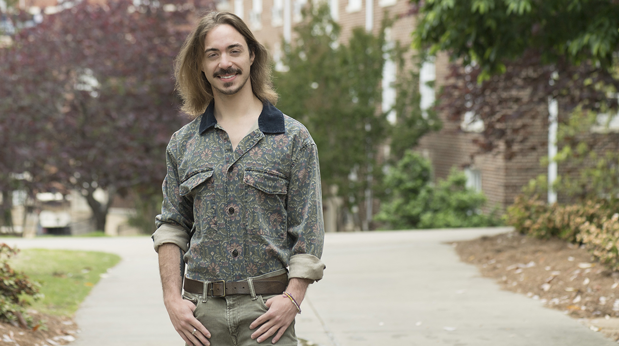 A young man stands with hsi hands in his pockets in front of a brick building.