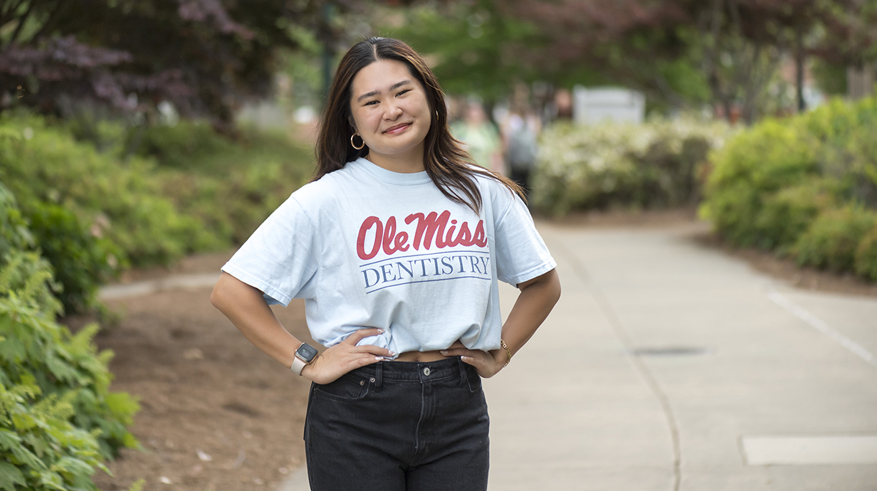 A young woman wearing a light blue Ole Miss T-shirt stands on a sidewalk in a park.