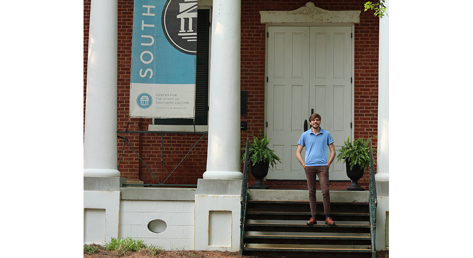 A young man stands on metal steps leading to a porch on a white-columned building.