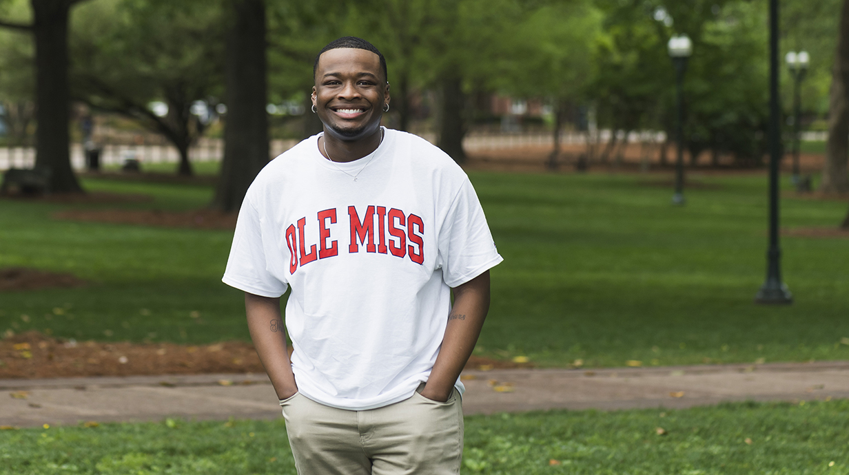A young man wearing a white Ole Miss T-shirt stands in a park.