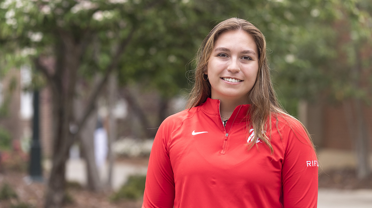 A young woman wearing a bright red rifle jersey stands in a park.