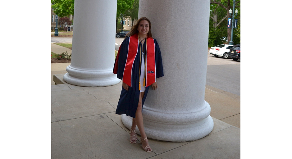A young woman wearing a graduation robe stands on a porch beside a white column.