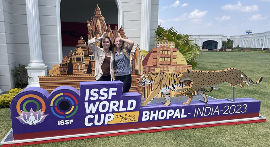 Two young women stand behind a sign for a shooting competition in India.