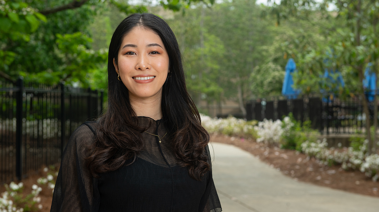 A young woman wearing a black dress stands near an iron fence in a park.