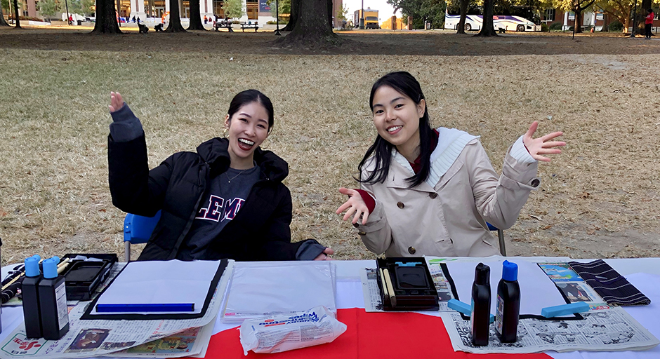 Two young women sit at a table lined with paper and printing materials in a park.