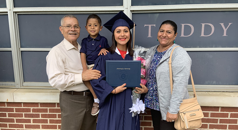 A family stands with a woman wearing graduation robes and a diploma folder.