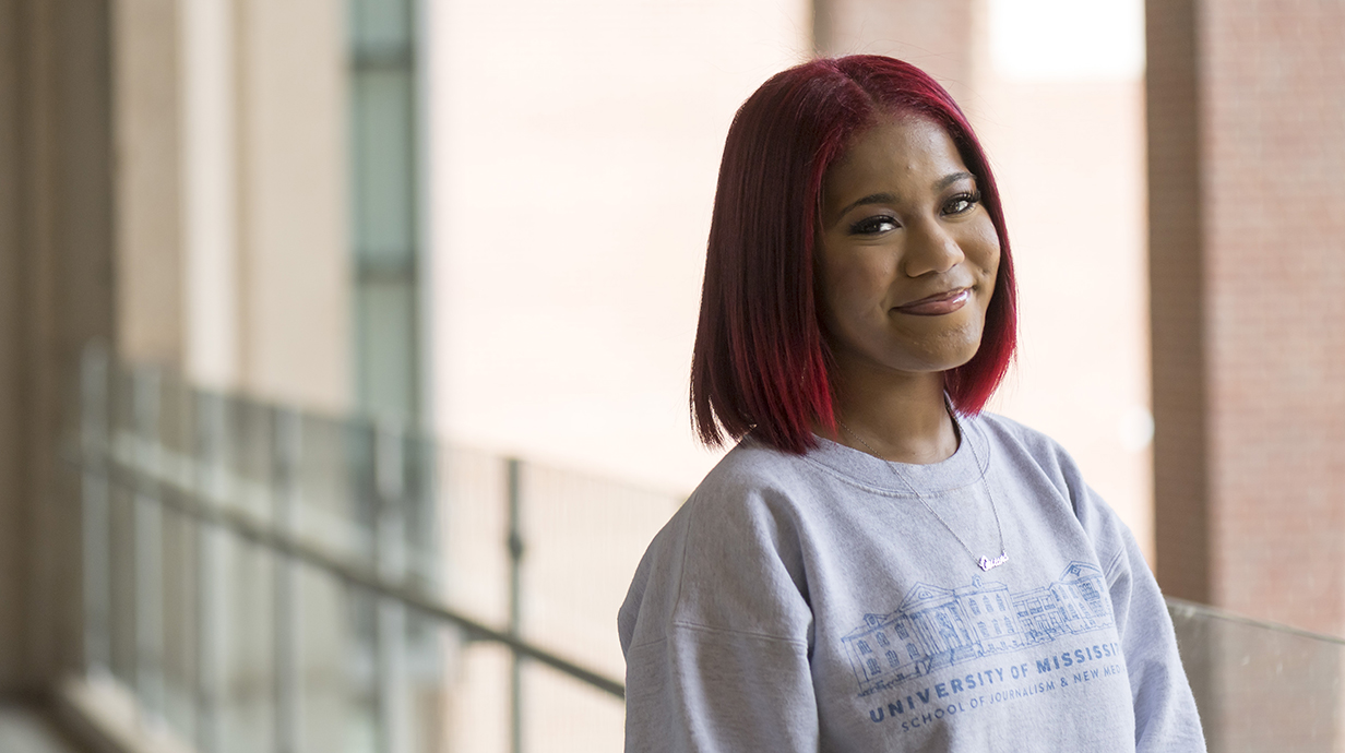A young woman wearing a School of Journalism sweatshirt stands on an outdoor balcony.
