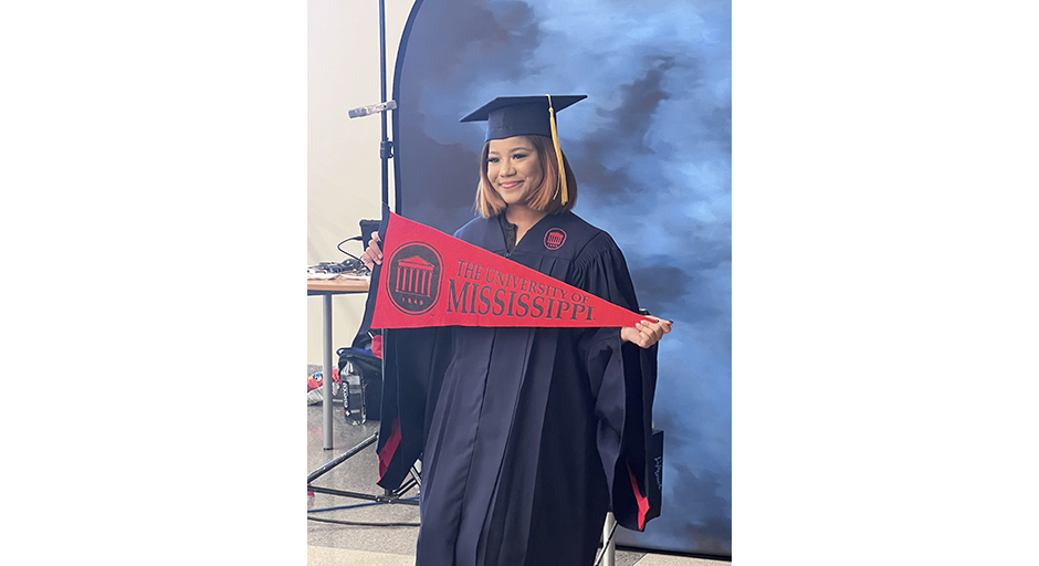 A young woman wearing graduation robes holds a red pennant reading University of  Mississippi.