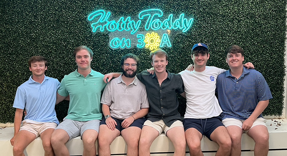 Six young men sit on a white bench in front of a neon sign reading "Hotty Toddy on 30A."
