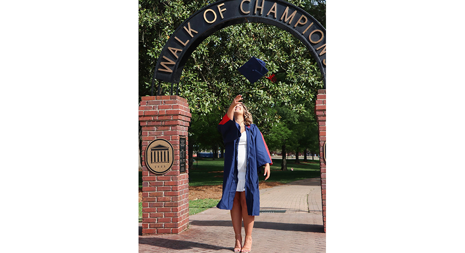 A young woman wearing graduation robes throws her hat into the air underneath an archway labeled as walk of Champions.