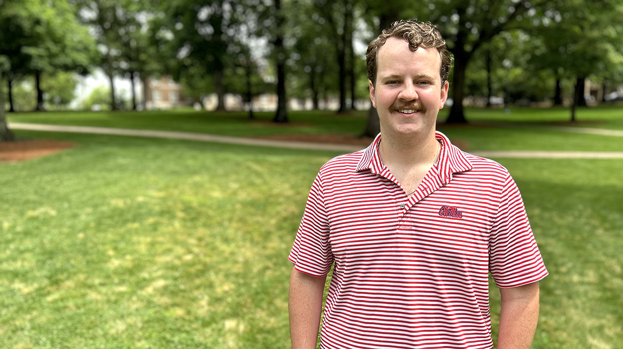 A young man wearing a red-and-white striped shirt stands in a tree-filled park.