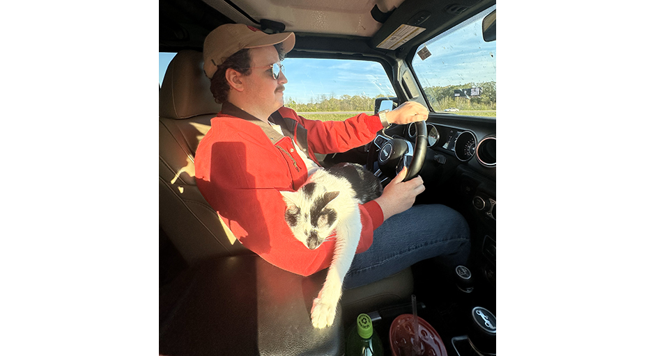 A young man drives a car with a cat in his lap.