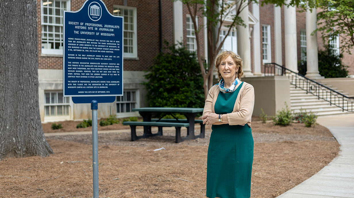 A woman stands next to a historical marker in front of a brick building.