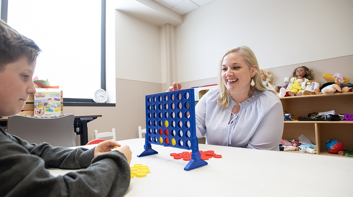 A woman and a young boy enjoy a game of Connect 4 in a playroom lined with toys.
