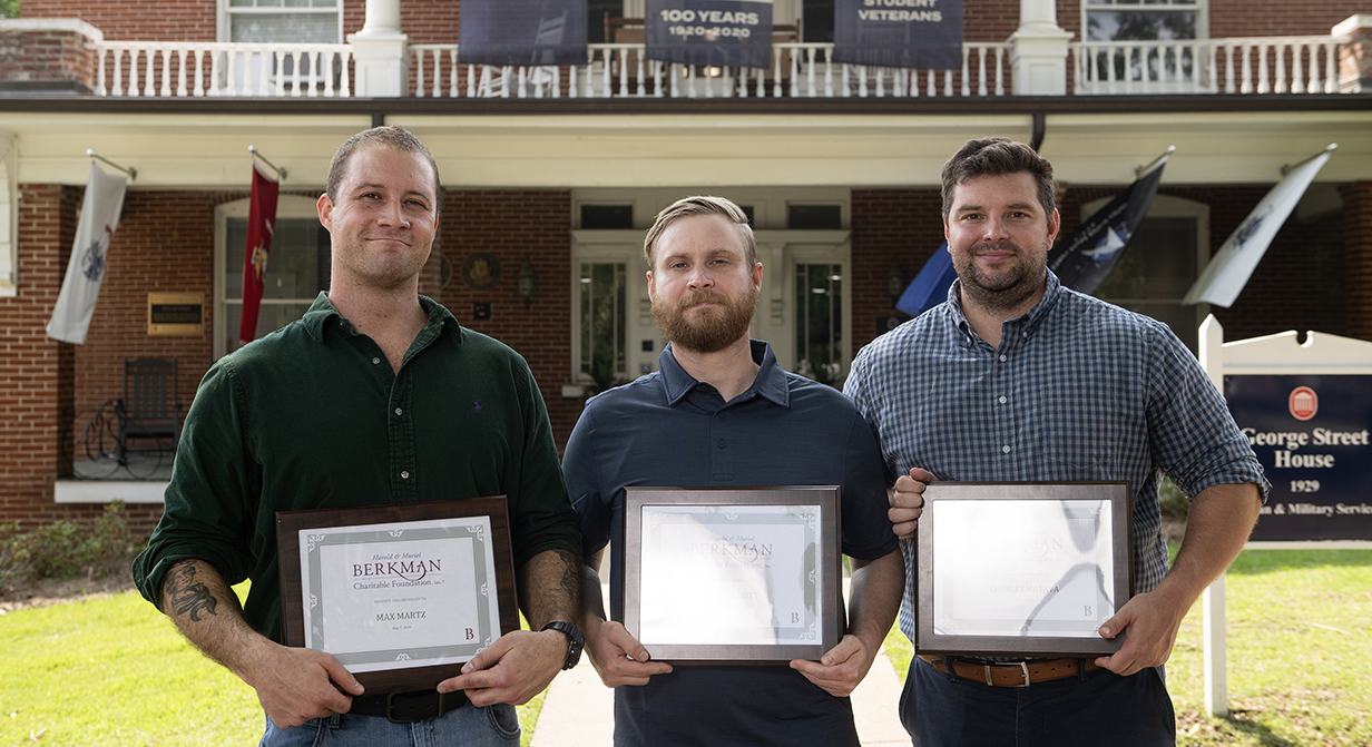 Three men stand outdoors holding award certificates.