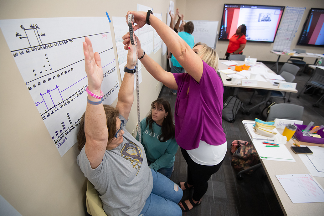A group of people participating in an educational activity inside a classroom. One woman, seated and wearing a gray shirt, raises her arms while another woman in a purple shirt measures her reach using a measuring tape. Charts with data and graphs are displayed on the walls, and various classroom materials and equipment are scattered on the tables. Several other participants are engaged in similar activities in the background, creating an active learning environment.