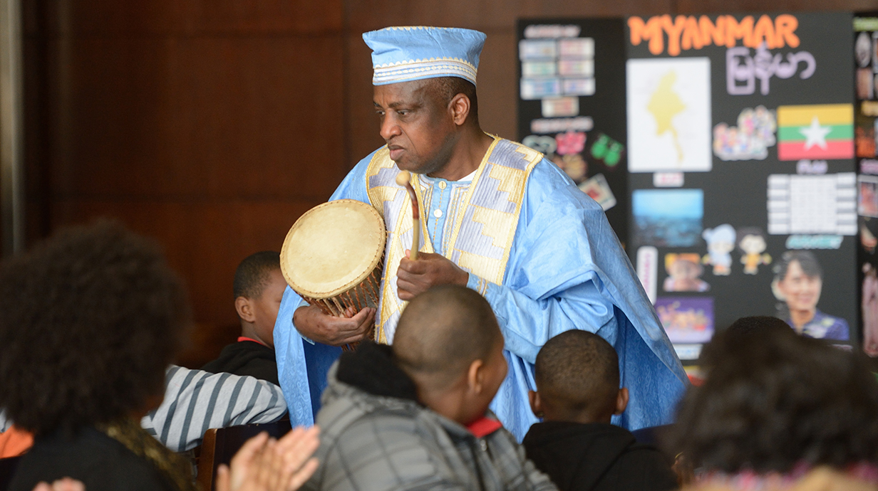 A man in a blue African robe plays a drum for a group of children in a classroom.