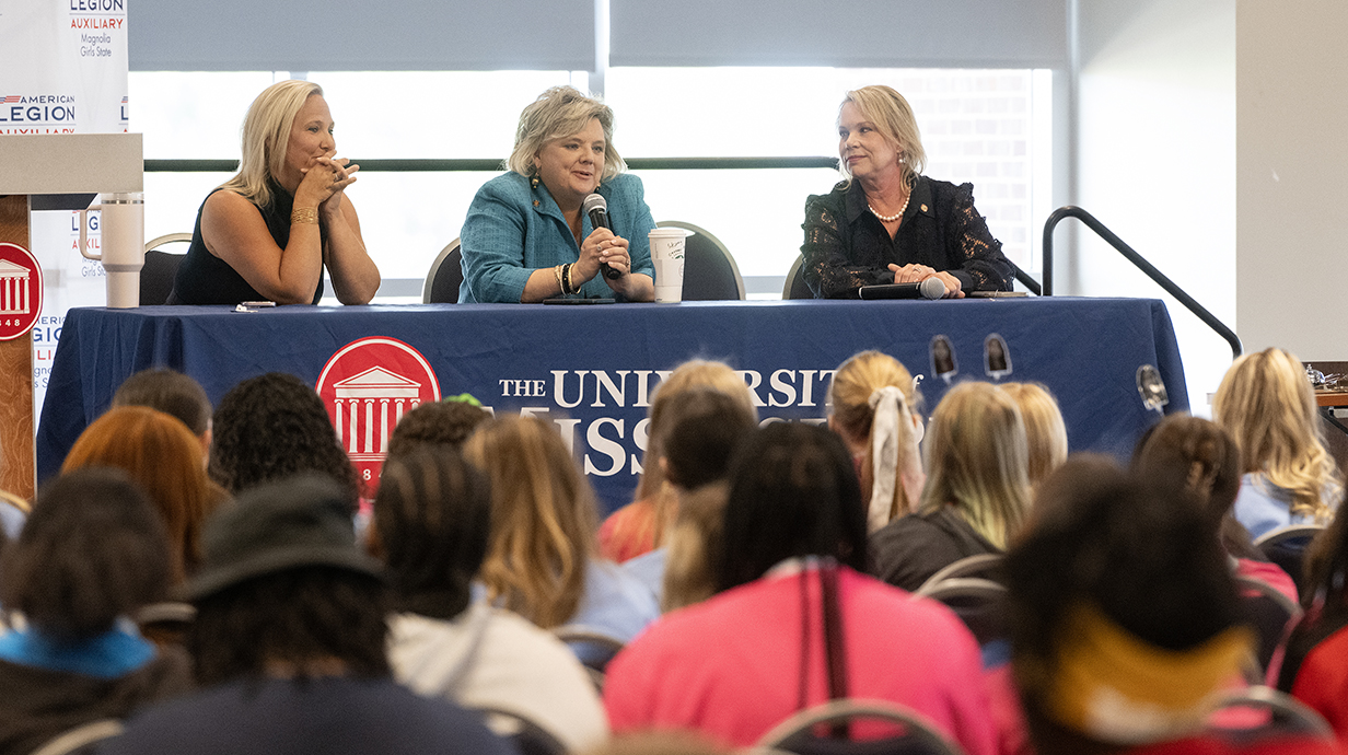 Three women sit behind a table on a stage in front of a conference hall full of young women.